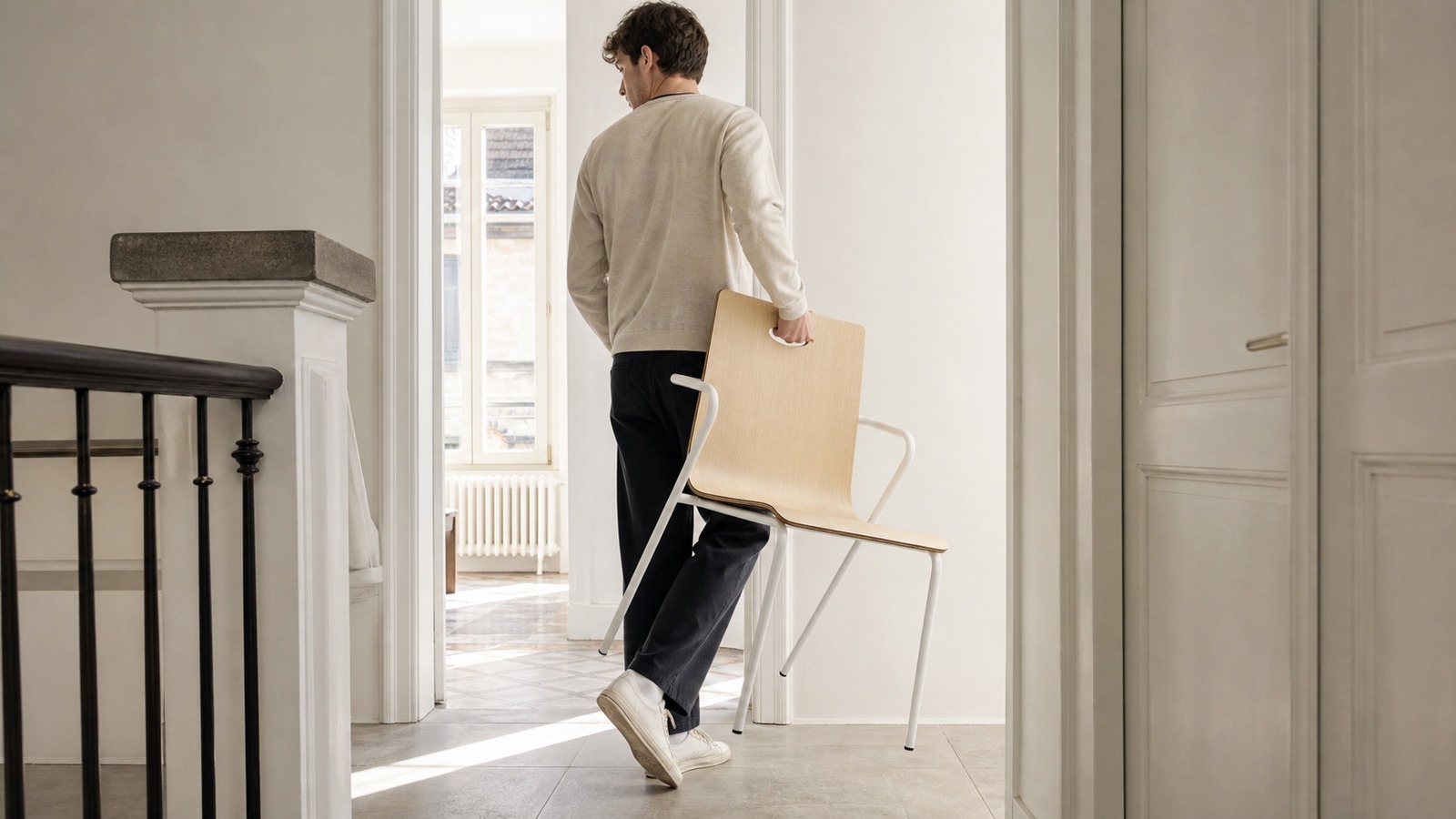 Urban renter carrying a lightweight bent plywood chair through an apartment corridor by its integrated handhold.